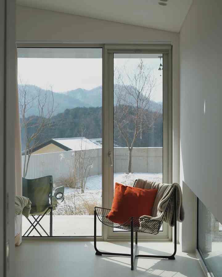 ホワイトトーンの寝室、大きなスライディング窓と黒の金属製アームチェア、オレンジのクッションとブランケットが置かれた温かみのある空間