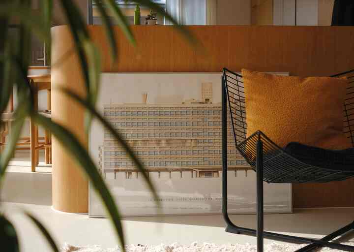 温かみのある木目調のリビングルーム、壁面に造り付けの収納とル・コルビュジエのポスターが置かれ、オレンジのクッション付きアームチェアが配置されたシンプルな空間