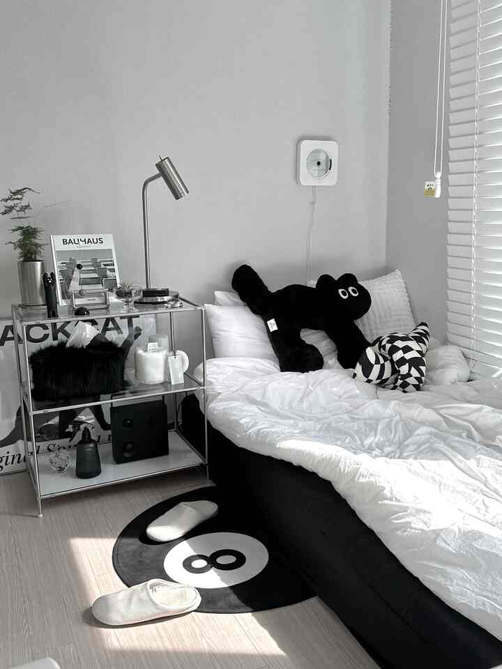 モノトーンの狭い寝室、黒い猫のぬいぐるみと8番のラグが置かれた黒いベッドとモジュラーシェルフが配置された実用的で整ったインテリア