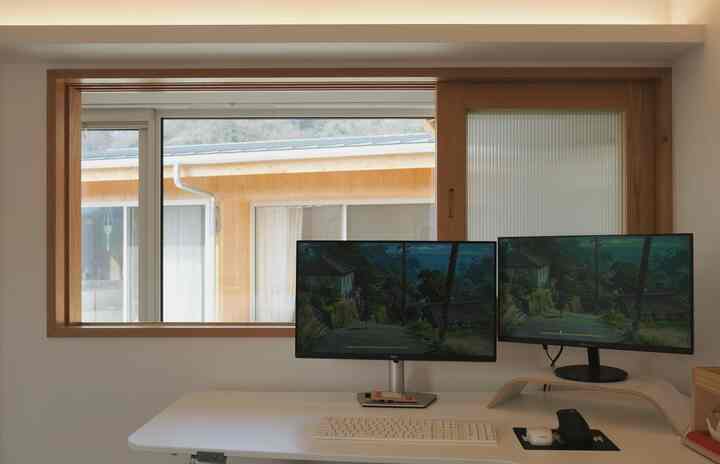 ウッドトーンの窓枠とホワイトのデスクが配置されたモダンなホームオフィス空間。デュアルモニターとキーボードがリモートワークに適している