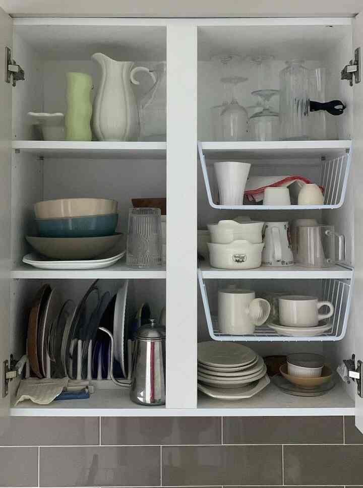 ホワイトとグレーのトーンのキッチンキャビネット内部、バスケットと皿整理ラックで整頓された食器とカップがある雰囲気のあるインテリア