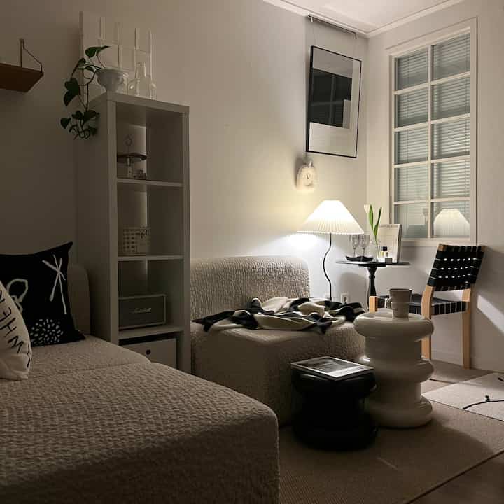 落ち着いたホワイトトーンのリビング、シンプルでミニマルな白いソファとスツール、温かみのあるフロアランプが心地よい雰囲気を作る空間