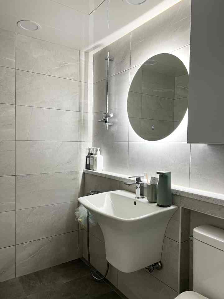 白とグレーのトーンによるコンパクトでモダンなバスルーム、丸型LED壁掛け鏡と機能的な洗面台が配置された清潔感のある空間