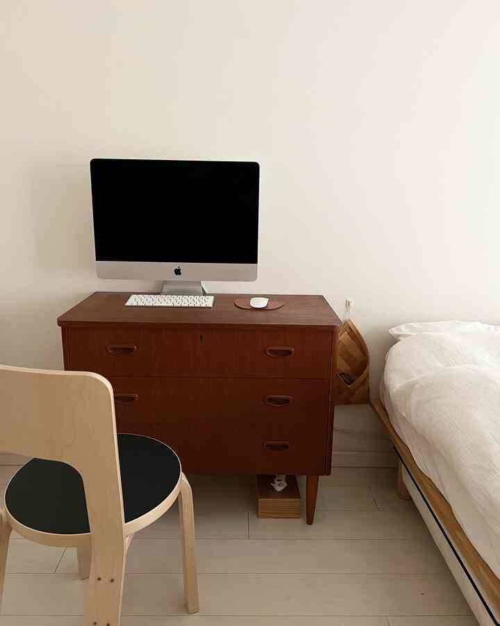 白い壁と寝具を背景にした寝室、中央にミッドセンチュリーモダンの木目調チェストをデスク代わりに使い、アルテックチェアが配置されたシンプルなホームオフィス空間