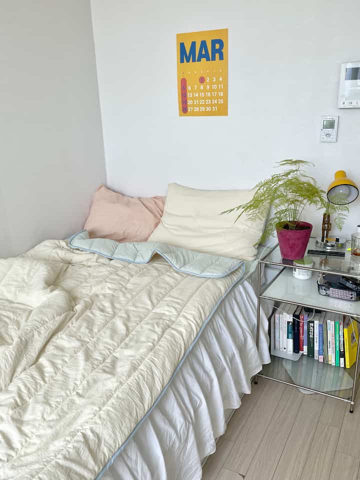 ナチュラルカラーとホワイトトーンの寝室、ベージュのベッドカバーと枕、ナイトテーブルの上に黄色いランプと緑の植物が配置された暖かみのある空間