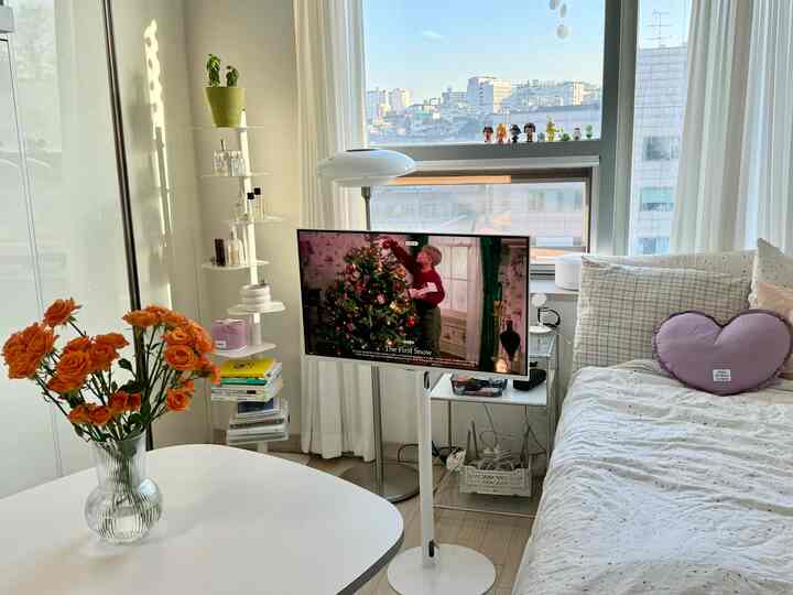 明るいホワイトトーンの寝室、右側にベッドと紫色のハートクッション、中央にテレビスタンドとフロアランプが配置された居心地の良い空間