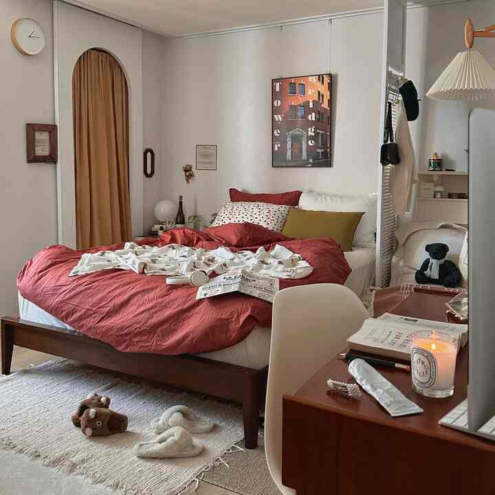 白い壁と茶色の木製家具が調和した二人用寝室、赤い寝具と居心地の良いデスクスペースがあるモダンでナチュラルな雰囲気のインテリア