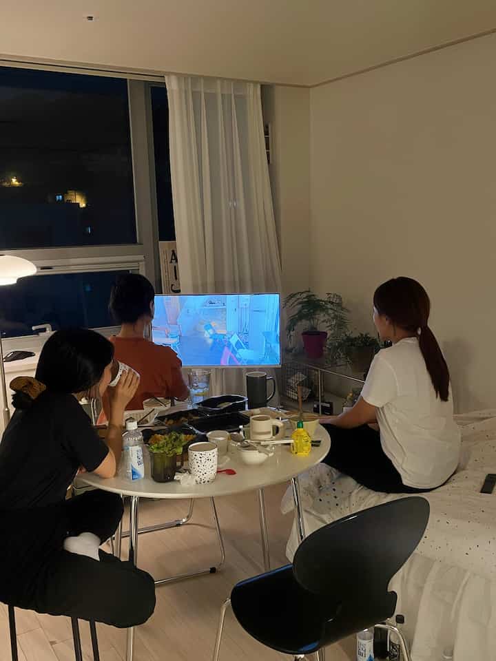 白と黒のトーンの寝室空間、丸いダイニングテーブルとチェアが配置され、3人が食事をしながらテレビを視聴する雰囲気のあるホームパーティーのインテリア