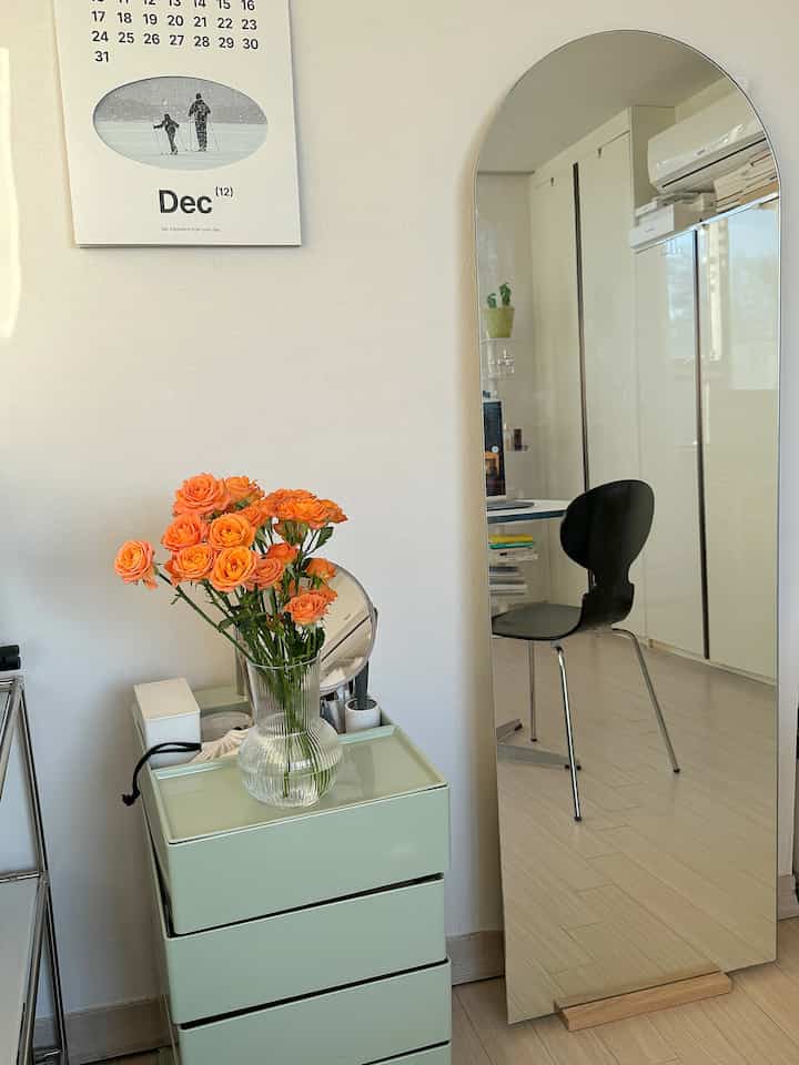 白い壁と明るい空間に、洗練されたミント色の収納チェストとオレンジ色の花瓶、大きなアーチ型の全身鏡が配置されたシンプルなパウダールーム