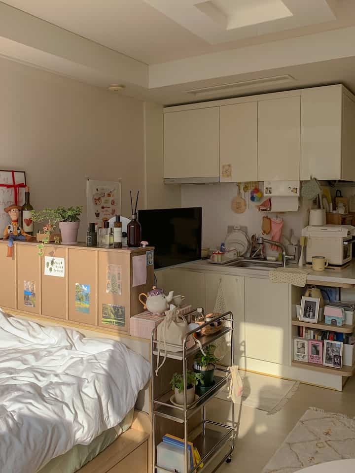 温かみのある木目調と白色のワンルーム、棚とチェストで寝室とキッチンが居心地よく仕切られた空間