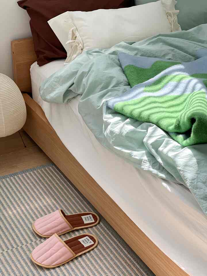 ウッドトーンのベッドがある寝室、ホワイトピローとミント色の掛け布団、ピンクのスリッパが置かれた温かみのある空間