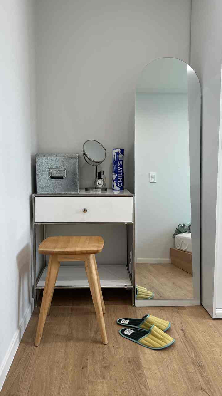 ナチュラルな木目調の床と壁、白いチェストと木製スツール、全身鏡が配置されたシンプルな化粧スペース