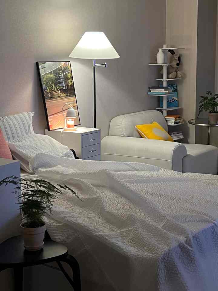 白とグレーの色調の寝室、ダブルベッドとあたたかいフロアランプ、ソファ、チェスト、植物が配置された雰囲気のあるインテリア