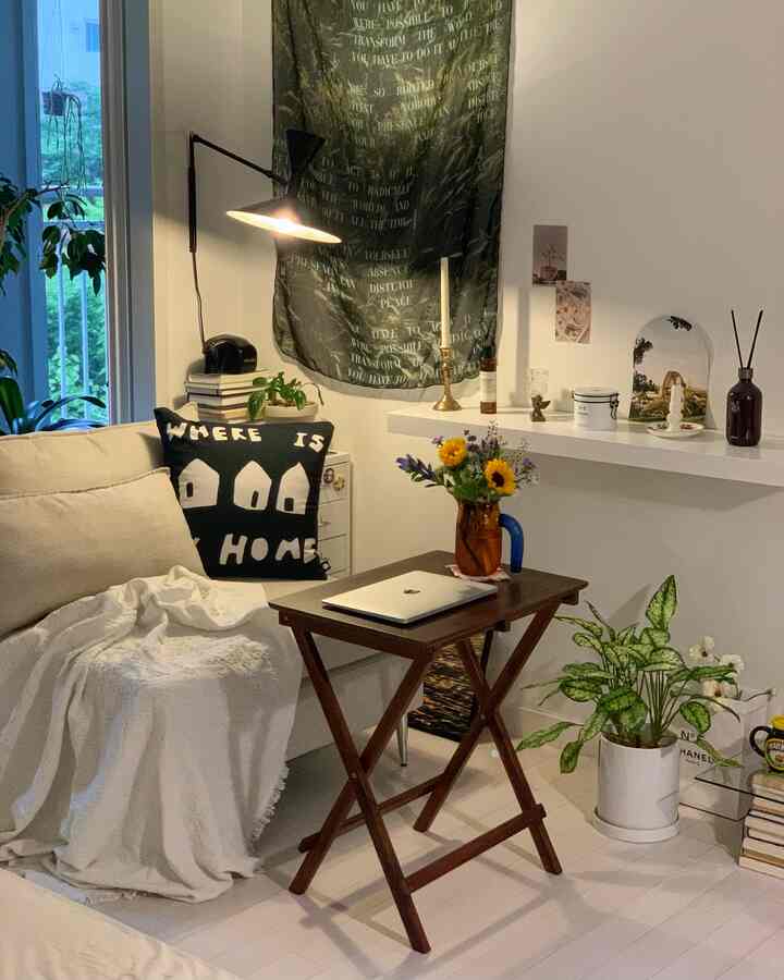 温かみのある木目調とベージュが調和する小さなリビングスペース、居心地の良いソファと折りたたみテーブル、自然な植物と花の装飾が配置された印象的な空間