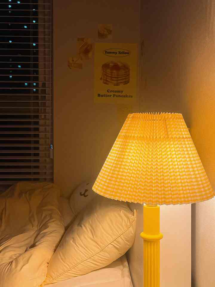 黄色の照明が灯る小さな寝室、クリーム色の寝具と柄のある黄色いテーブルランプが温かみのある雰囲気を演出する空間