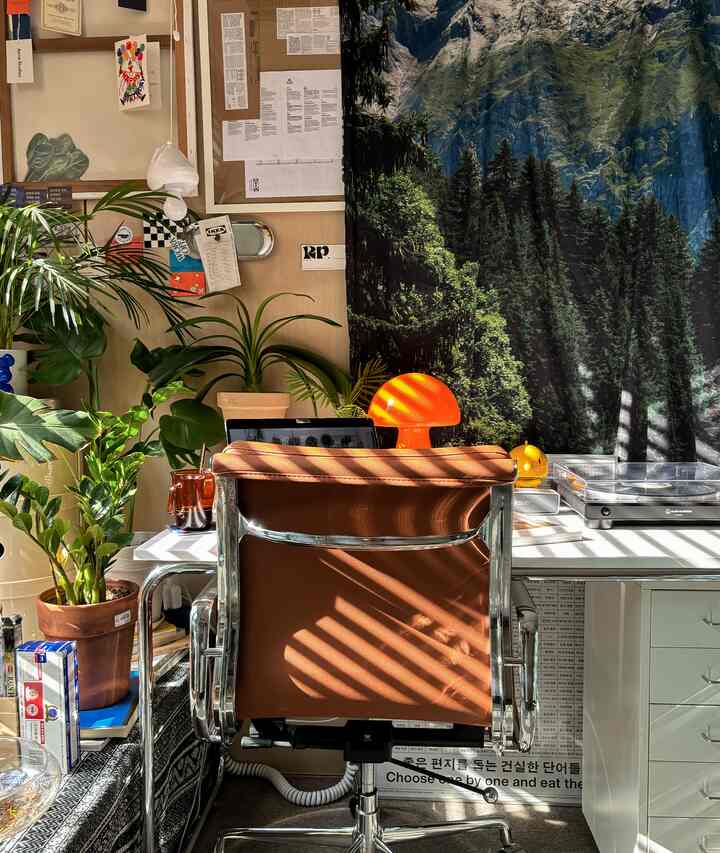 ブラウンのレザー椅子と緑の植物が配置されたホームオフィス、壁には自然の風景を描いたタペストリーと額縁が飾られた温かみのある空間