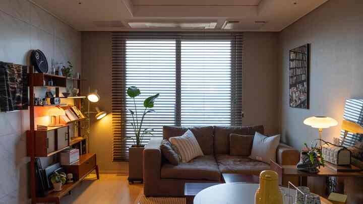 ブラウンの木製家具とナチュラルなトーンのリビング、広いソファとウッドブラインド、複数の照明と観葉植物が配置された雰囲気のあるインテリア