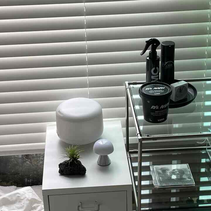 ホワイトトーンのブラインドとメタルモジュラーシェルフがある寝室、ナイトテーブルに植物とムードランプが調和した清潔感のある空間