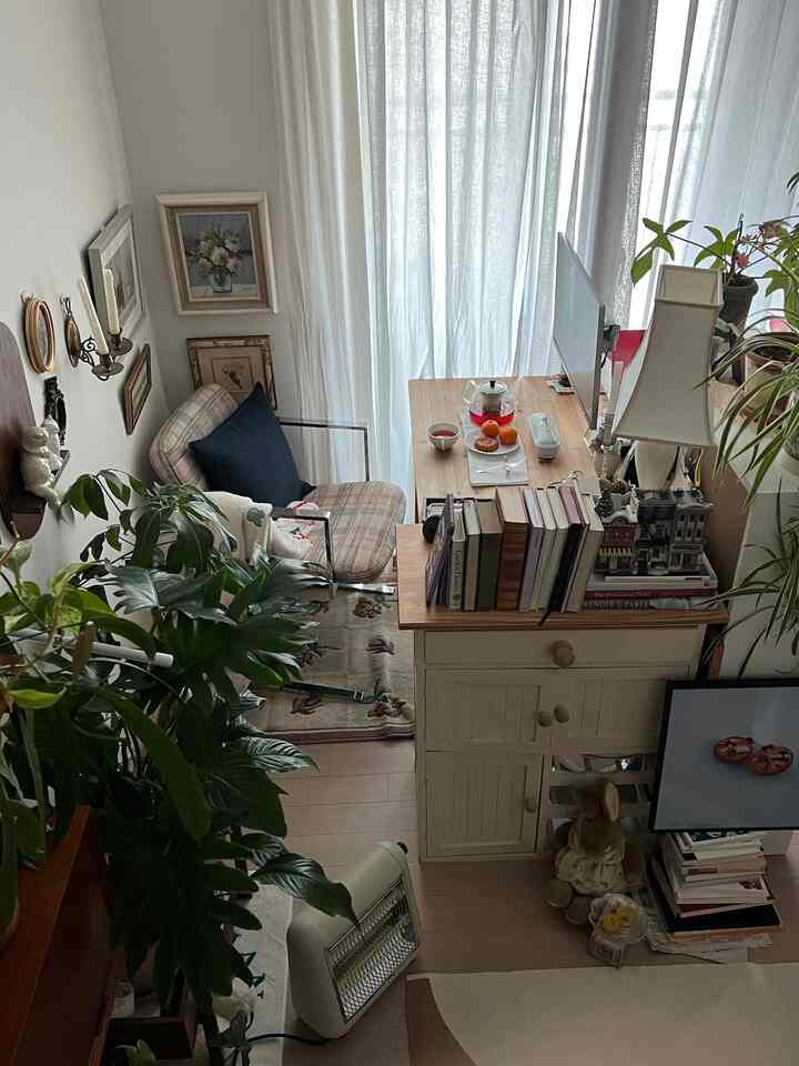 明るいアイボリーとナチュラルウッドのトーンのホームオフィス、机や本棚、ラグ、カーテン、額縁、植物が配置された落ち着いた読書と在宅勤務の空間