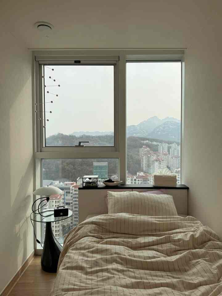 ベージュとホワイトのトーンの小さな寝室、中央にシングルベッドと黒のナイトテーブルが配置され、窓越しに街と山の景色が見える印象的な空間