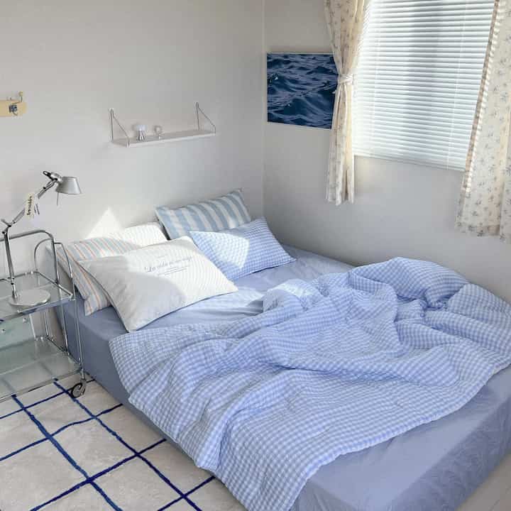 白い壁と青いチェック柄の寝具がある6畳の一人暮らし用の寝室、自然光が柔らかく差し込む空間
