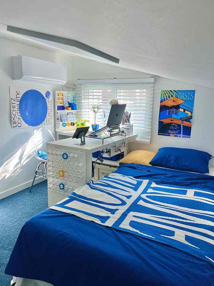 青と白の色調が映えるコンパクトな寝室、傾斜天井の下にベッドとホームオフィスが配置された落ち着いたインテリア