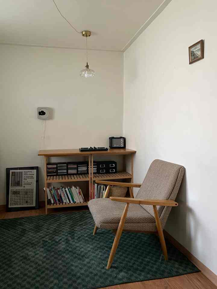 ナチュラルな色調の小さな寝室コーナー、木製の棚とアームチェア、緑のラグが配置された雰囲気のあるインテリア