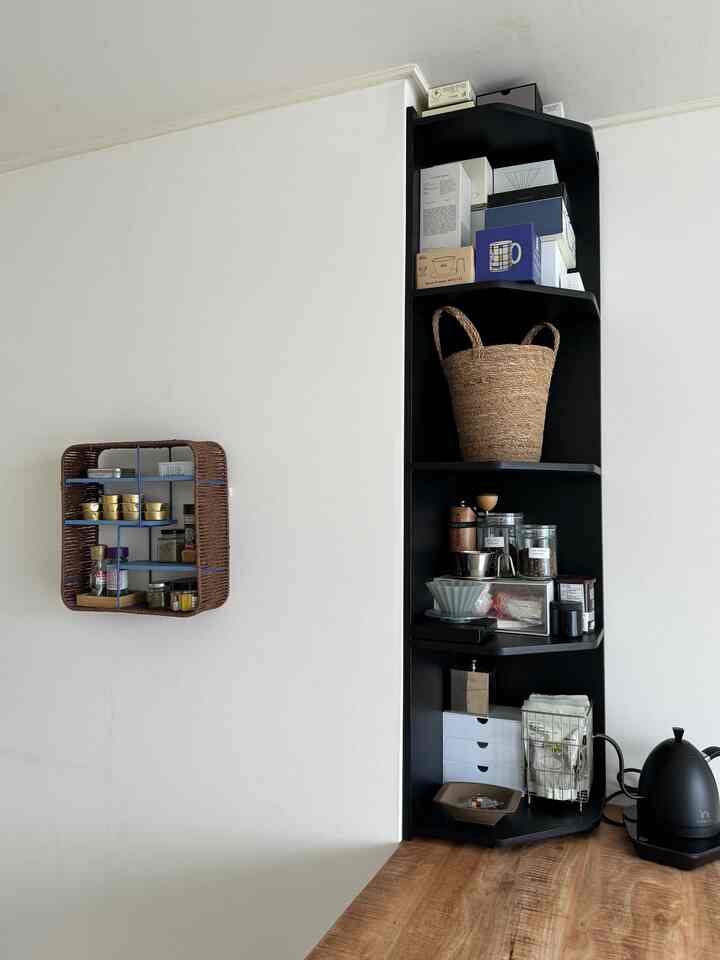 ナチュラルカラーの木製テーブルと黒のコーナー棚が配置されたキッチンスペース、整頓されたコーヒー器具が印象的な清潔かつ機能的な雰囲気