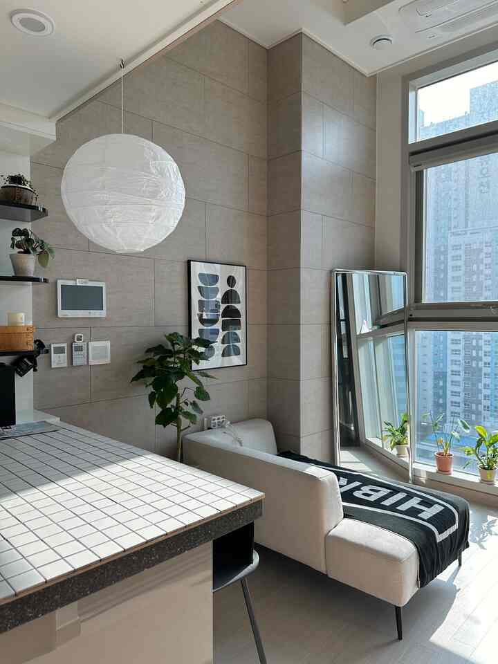 ベージュとホワイトの色調のリビング、自然光差し込む大きな窓のそばにソファ、壁アート、鏡、植物が配置された雰囲気のあるインテリア