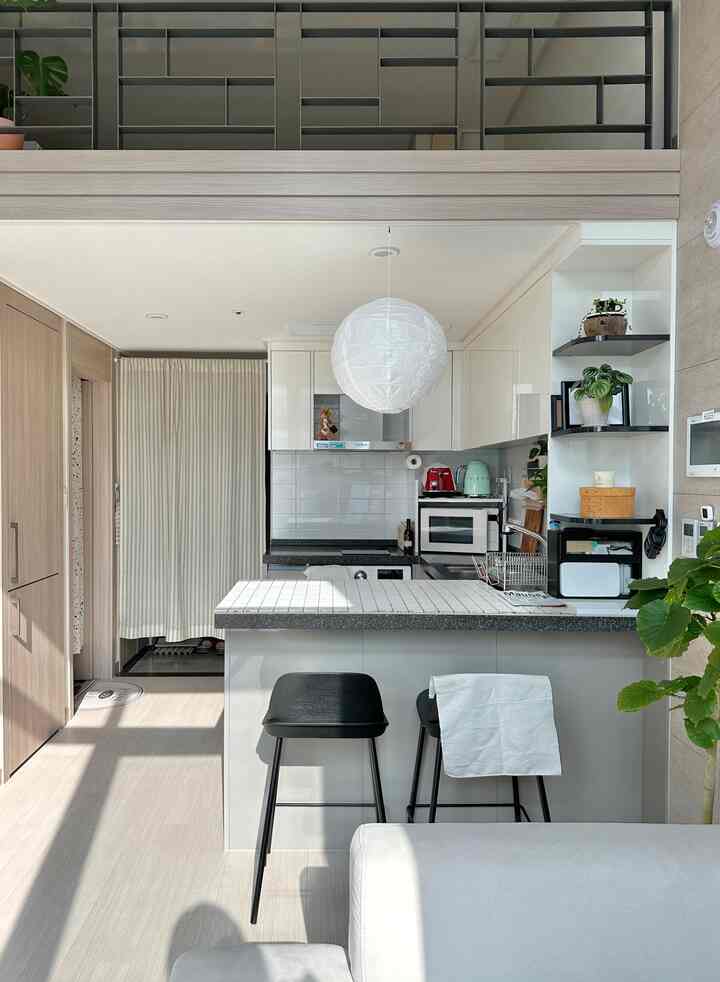 ホワイトとブラウンのモダンなキッチン空間、中央にペンダントライトとバースツール、左側にカーテンと玄関が見えるすっきりとしたインテリア