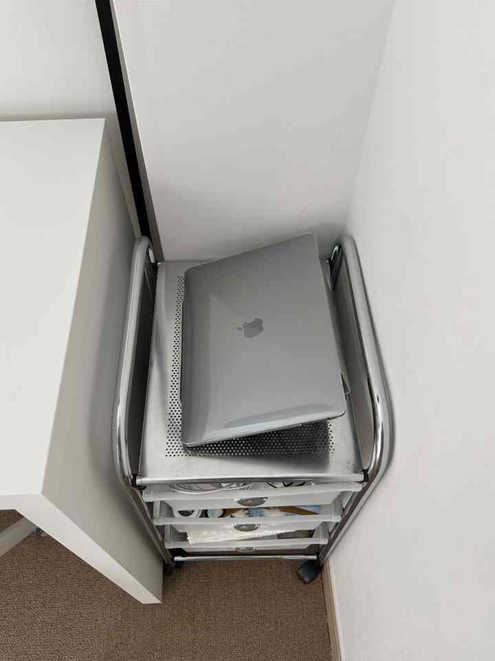 白い壁と机の隣に配置されたクローム製引き出しトロリーと、その上に置かれたグレーのアップル・マックブックがあるコンパクトなホームオフィス空間