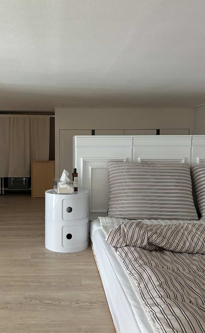 ナチュラルなブラウンの床と白いナイトテーブル、ストライプの寝具が配置された落ち着いた寝室のインテリア