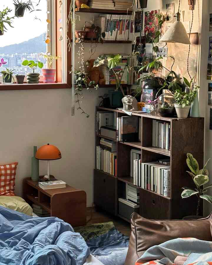 木目調のナチュラルな寝室空間、本棚とオレンジのテーブルランプ、多彩な植物が配置された雰囲気のあるインテリア