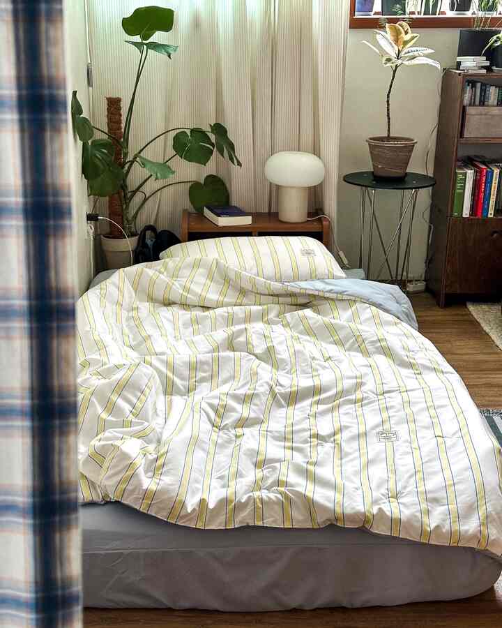 明るいイエローストライプの寝具があるナチュラルな寝室、木目調の家具と植物が温かみのある雰囲気を演出する空間