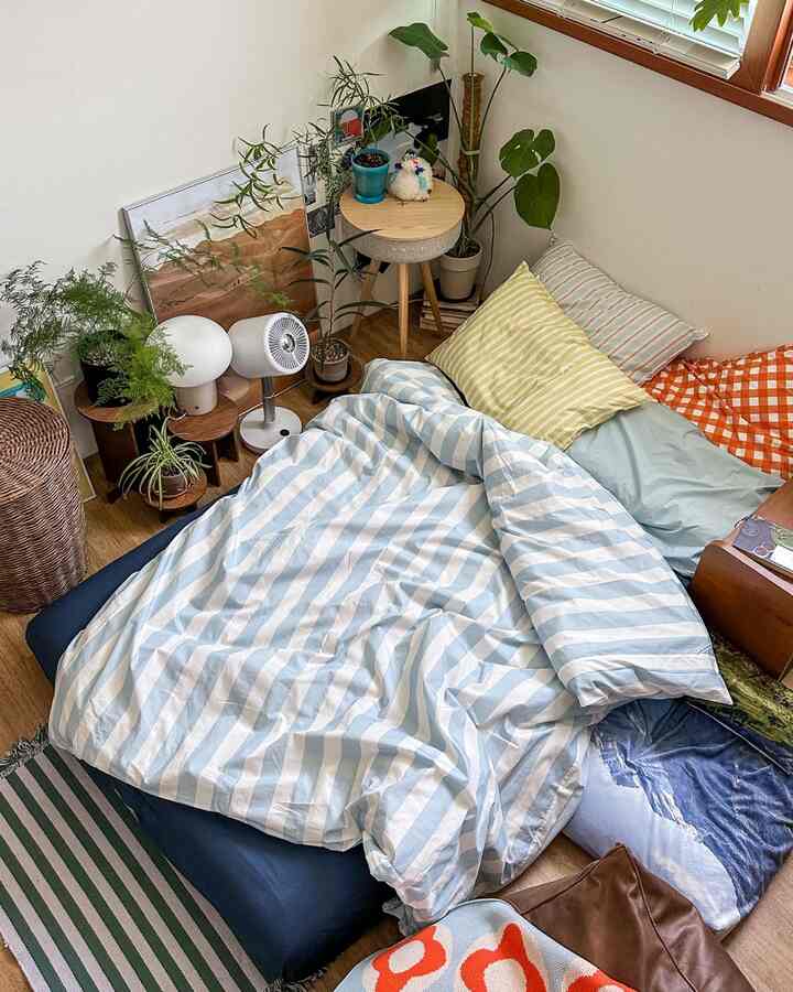 ナチュラルな木目調の床と複数の枕があるブルーストライプの寝具を配した狭い寝室、植物とスツールが温かみのある雰囲気を演出