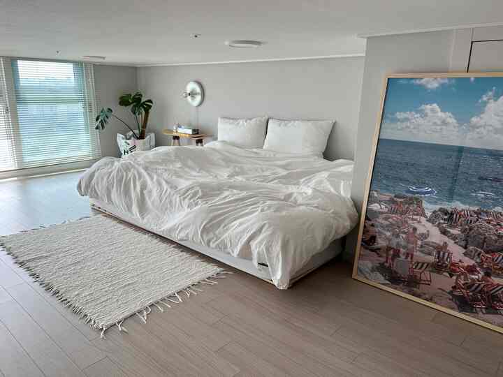 ホワイトモノトーンの寝室、広い窓にブラインド、白い寝具とラグ、ナチュラルウッドの床が配置された雰囲気のあるインテリア