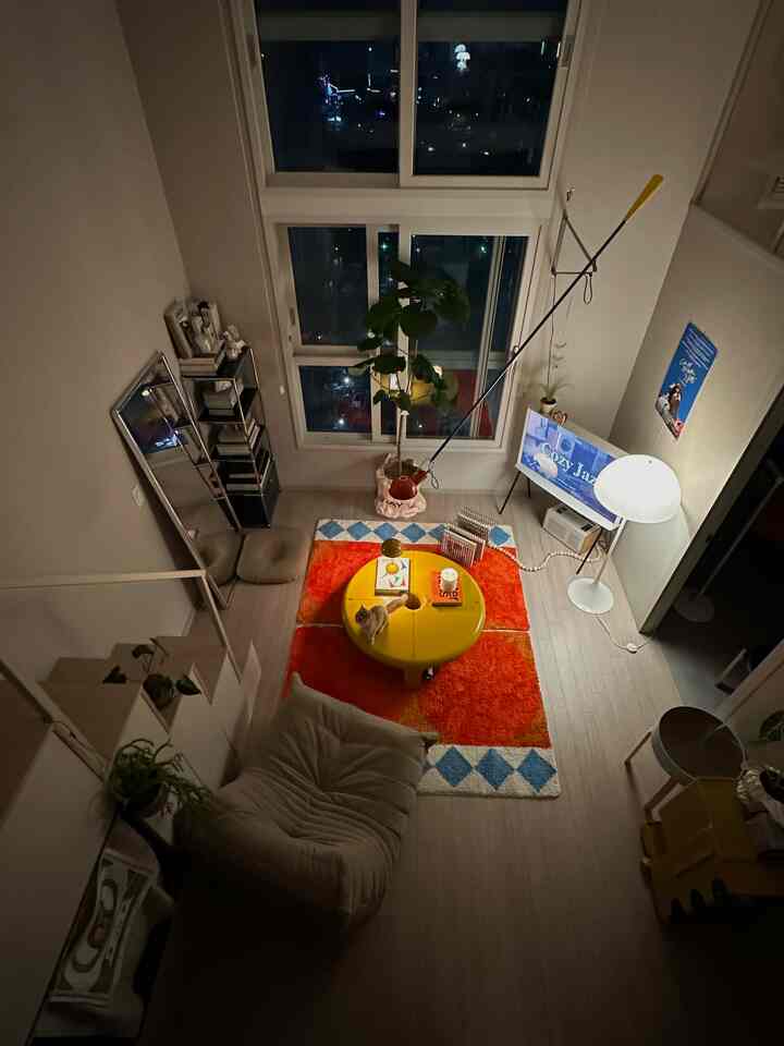 ベージュとナチュラルカラーのリビングルーム、中央に黄色い丸テーブルと赤のラグが配された居心地の良い空間