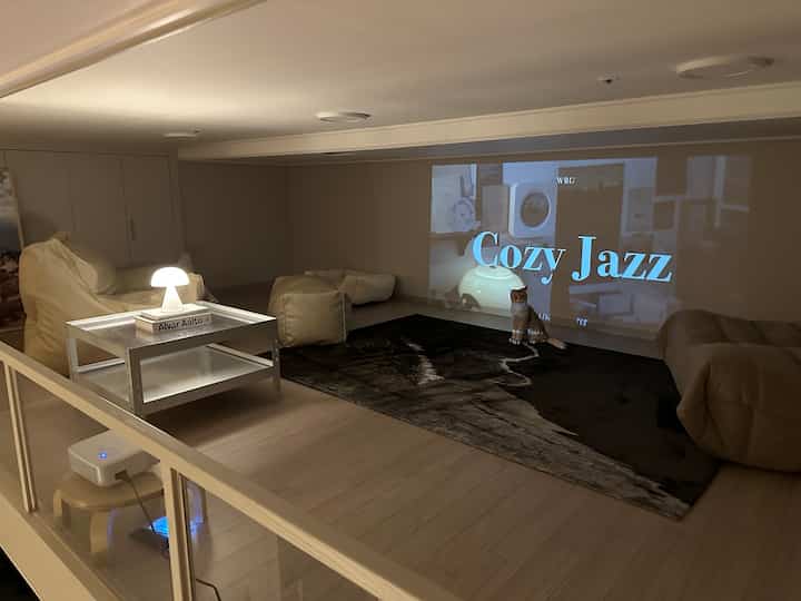 ナチュラルカラーの小さなホームシネマ空間、クリーム色のビーズクッションとダークラグ、プロジェクターとスクリーンが配置された雰囲気のあるリラックススペース