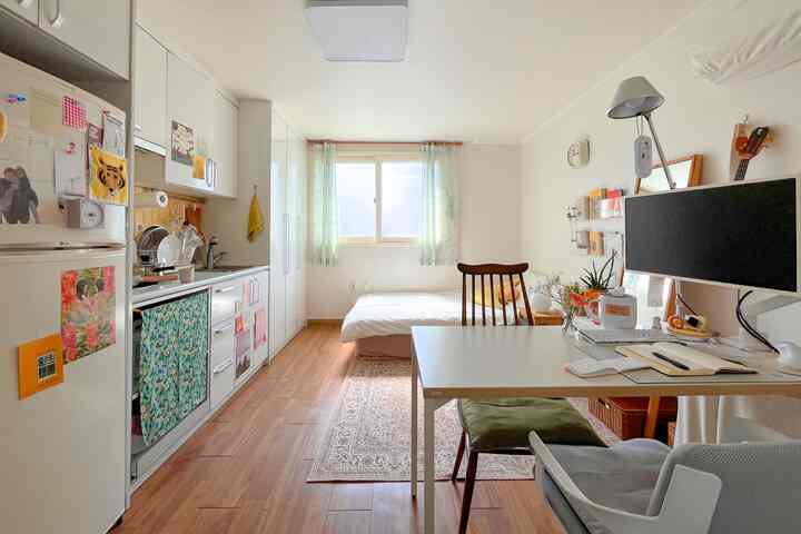 白とナチュラルカラーの調和が取れたワンルーム空間、窓際のベッドと作業用デスク、キッチン設備が配置された明るく居心地の良いインテリア