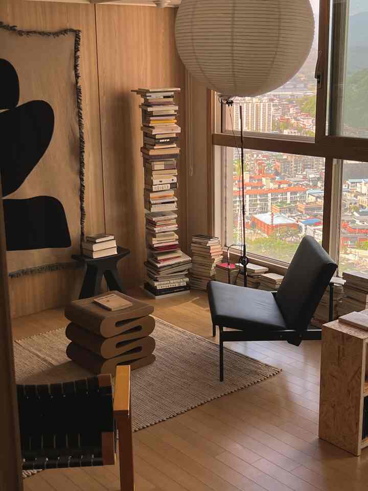 明るい木目調の壁と床、黒のアームチェアと積み重ねられた本が配置された居心地の良いワンルームのリビング