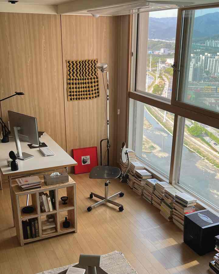 ベージュの木目調が特徴的なホームオフィス、窓際にデスクとオフィスチェアが配置され、本が積まれた雰囲気のある書斎