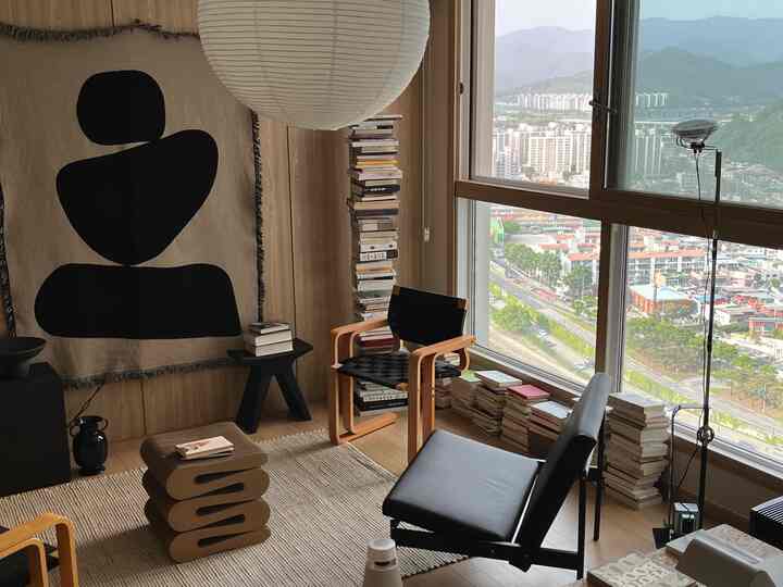 ナチュラルモダンのリビングルーム。大きな窓から自然光が差し込み、スツールやアームチェア、ラグ、積まれた本が配置された落ち着いた印象の空間。