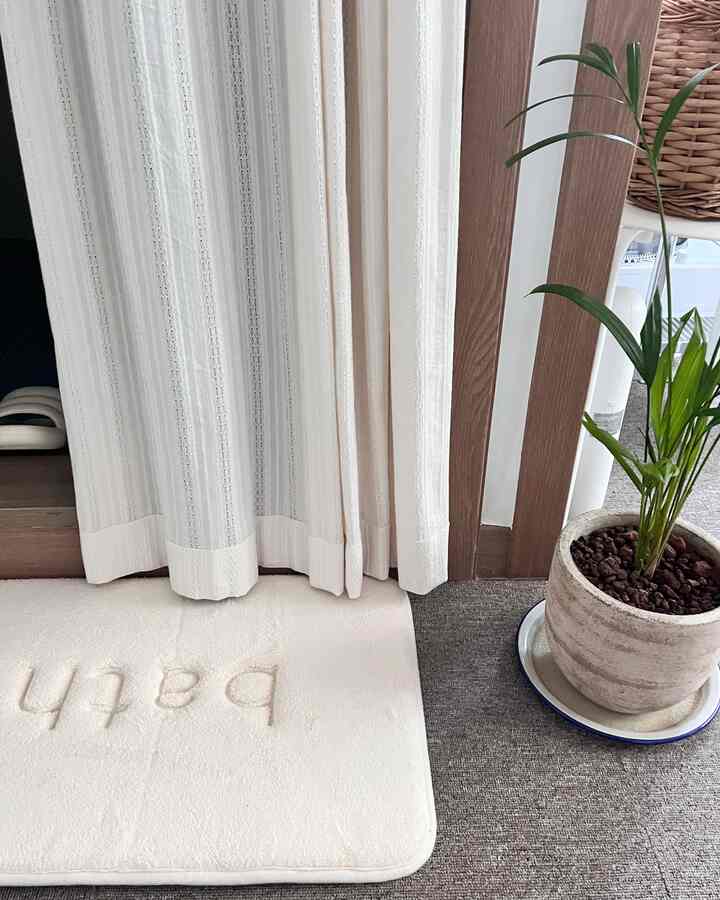 ベージュのバスマットと白いレースカーテンが配置された自然な色調のトイレ入口の雰囲気のあるインテリア