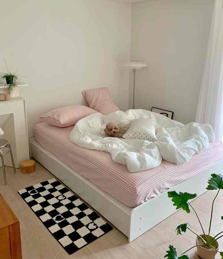 白とピンクのトーンの寝室、中央にクッションと布団のあるベッド、ペットの犬がくつろぎ、フロアランプと植物が配置された落ち着いた空間