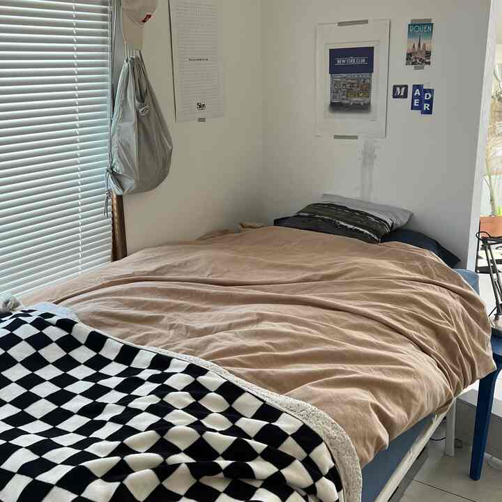 ベージュの掛け布団とチェッカーブランケットが置かれた狭い一人暮らしの寝室、窓の横にカーテンが設置された空間