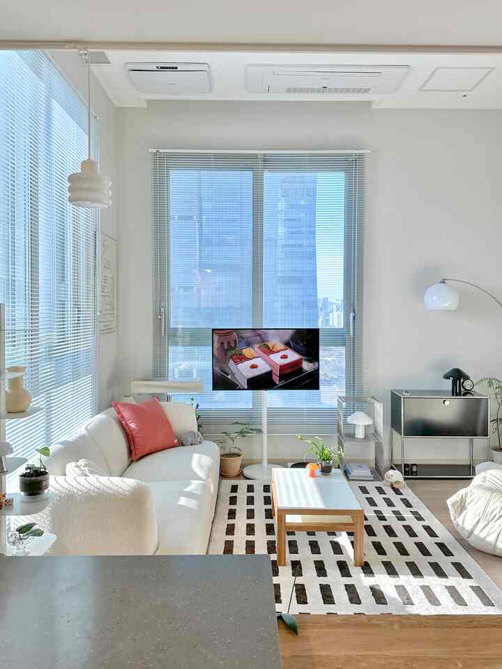 白とライトウッドトーンの調和したリビング、モダンなソファとラグ、ブラインド付きの窓が明るく清潔感のあるインテリア