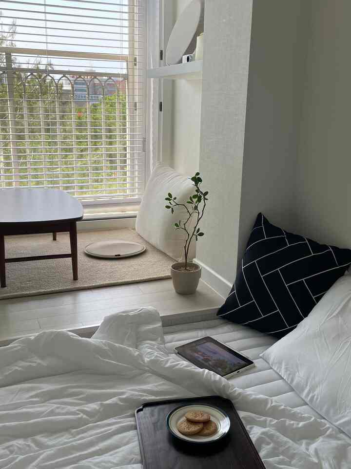 ナチュラルな色調の寝室、木製テーブルと平床、大きな窓に木製ブラインドが調和した雰囲気のある空間