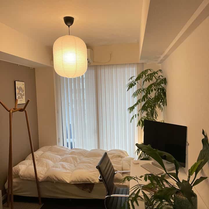 白と茶色のトーンが特徴のワンルーム寝室兼ホームオフィス、植物とコートラックが配置された自然でモダンな空間