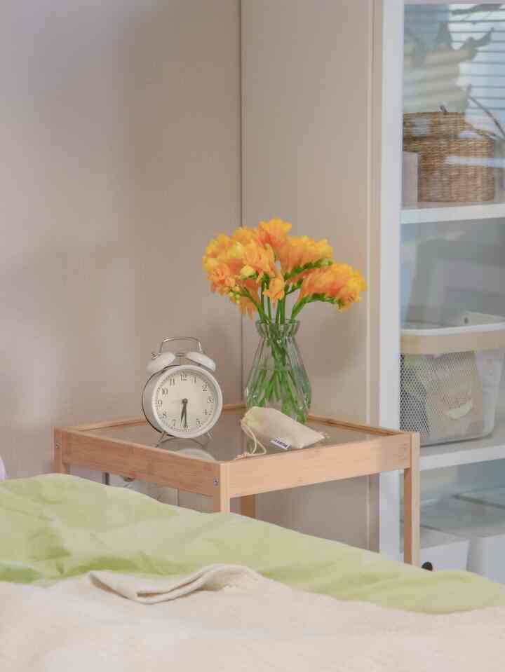 ナチュラルカラーの寝室、木製ナイトテーブルの上にアラーム時計と黄色のフリージアの花瓶が配置された落ち着いた空間