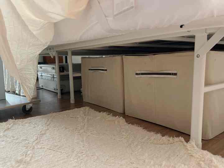 白を基調としたワンルームの寝室、高さのある金属製ベッドフレームの下にベージュの収納ボックスとスーツケースが整頓された印象的な収納スペース